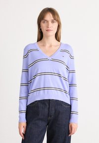 Pull violet clair avec un col en V, présentant des rayures horizontales noires et blanches. Tissu doux et coupe décontractée, associé à un jeans en denim foncé.