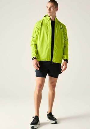 Jonge man in neon groene jas, zwarte korte broek en zwarte sneakers, staand tegen een effen lichte achtergrond en kijkt naar rechts.
