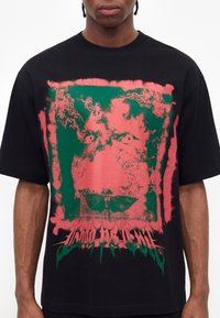 Άτομο φοράει μαύρο oversized t-shirt με αφηρημένο πράσινο και κοραλλί γραφικό που μοιάζει με παραμορφωμένο πρόσωπο και κείμενο στο κάτω μέρος.