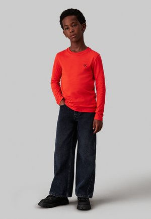 Camicia a maniche lunghe di un brillante colore rosso con scollo rotondo e piccolo logo; abbinata a jeans larghi blu scuro e scarpe nere.