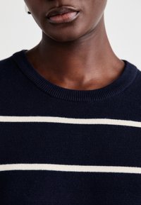 Tumesinine kudumiga sviiter, millel on ümmargune kaelus ja kaks valget horisontaalset triipu rinna kohal. Smooth tekstuur ja soonikkoes mansetid.