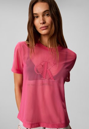 Νεαρή γυναίκα φοράει διάφανο ροζ μπλουζάκι Calvin Klein πάνω από αθλητικό σουτιέν με λογότυπο, στέκεται μπροστά σε γκρι φόντο.