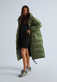 Garšs olīvu zaļš pūkains mētelis ar kapuci, knopku aizdari un sānu kabatām, apvienots ar melnu pieguļošu krekli un melnām motokrosa šortiem.