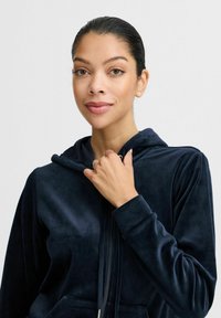 Navyblå velour zip-up hoodie med huva, långa ärmar och kenguruficka. Slät textur och metallisk dragkedjedetalj.