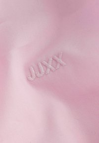 Lys rosa stoff med en glatt tekstur har brodert tekst "JUXX" i matchende trådfarge, som er litt hevet mot bakgrunnen.