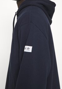 Námořnická modrá mikina s kapucí na šňůrku, měkká textura a bílý štítek Adidas Adventure na levém rukávu. Klasický design.