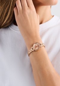 Roosa kuldne kell ümmarguse kellanäidiku, link-rihma ja tekstureeritud äärisega. Kellanäidikul on minimaalsed aktsendid ja peen disain.