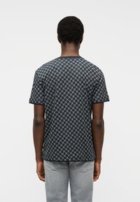 Άνδρας με κοντά σγουρά μαλλιά, φοράει μαύρο πουκάμισο με σχέδια και κοντά μανίκια και ανοιχτό γκρι τζιν, στέκεται με την πλάτη προς τα εμπρός σε λευκό φόντο.