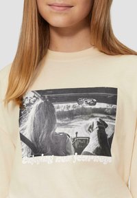 Cremefarbener Sweatshirt mit einem schwarz-weißen Grafikmotiv eines Menschen und eines Hundes im Auto, darunter der Text "Neue Reisen genießen".