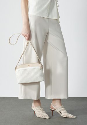 Femme tenant un sac à main blanc texturé, portant un pantalon beige à jambes larges et des escarpins slingback tissés beiges sur un sol gris.