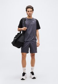 Νεαρός άνδρας με αθλητικό ντύσιμο σε μαύρο και γκρι, στέκεται κρατώντας μια μαύρη τσάντα γυμναστηρίου στον ώμο του, μπροστά σε καθαρό λευκό φόντο.