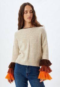 Maglione lavorato a maglia beige con una vestibilità ampia, presenta una tasca sul davanti e maniche con ponpon colorati in marrone, arancione e giallo.