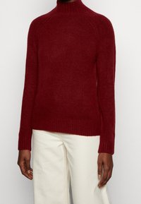 Viininpunainen neulepusero korkealla kauluksella, ribbireunuksilla hihoissa ja helmassa, pehmeällä tekstuurilla ja sopivalla siluetilla. Yhdistetty valkoisiin housuihin.