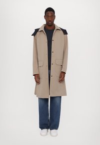 Bežna trench coat z temnimi ramenskimi poudarki, dvema sprednjima žepoma, gumbi za zapiranje in sproščenim krojem, oblečena čez mornarsko modro majico in kavbojke.