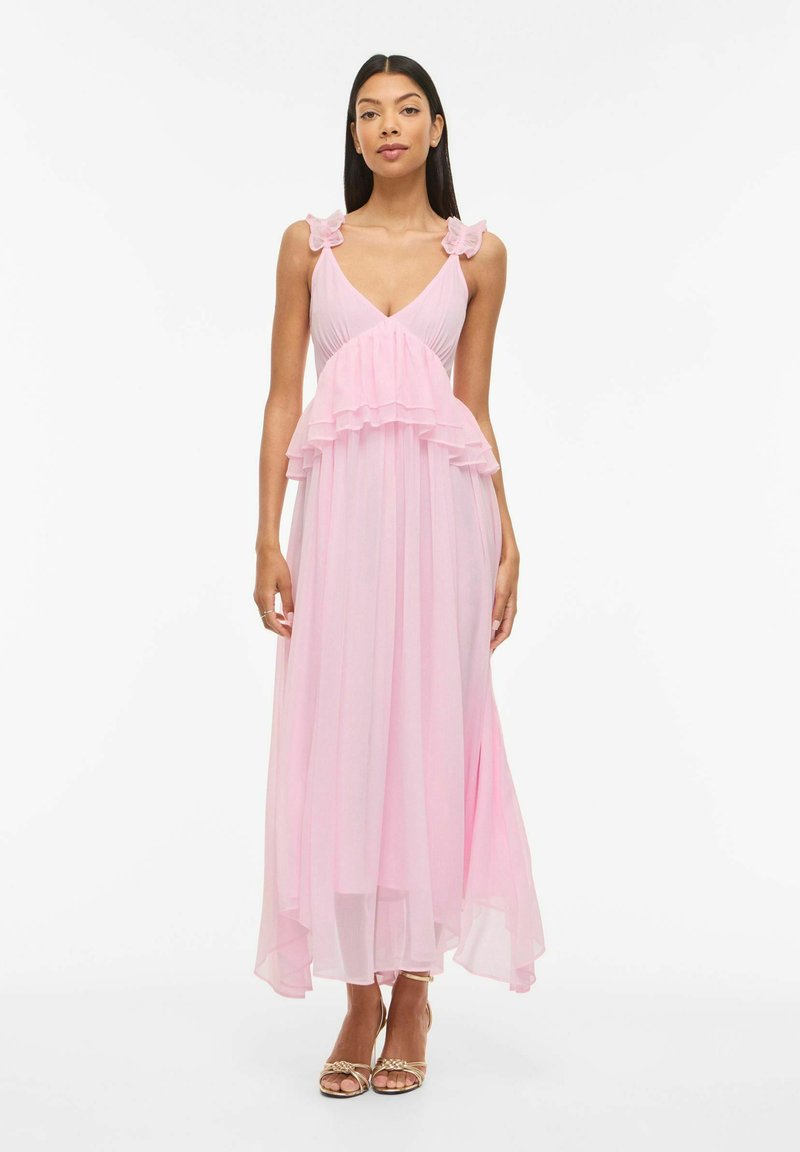 Femme debout portant une robe longue rose sans manches avec des volants sur les bretelles et le corsage, assortie à des talons hauts dorés à lanières.