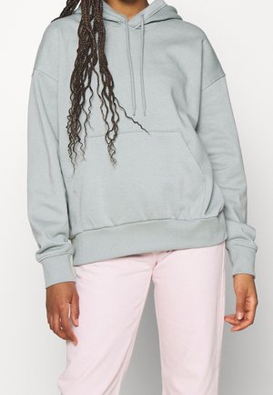 Personne portant un sweat à capuche gris clair avec poche avant et pantalon rose, debout devant un fond clair uni.