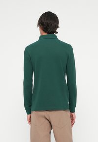 Man met donker haar in een donkergroen poloshirt met lange mouwen en beige broek, staand met de rug naar de camera tegen een witte achtergrond.