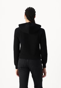 Haut à capuche noir côtelé avec une capuche proéminente, des manches ajustées et un ourlet court. Associé à un jean foncé, mettant en valeur un design en tricot texturé.