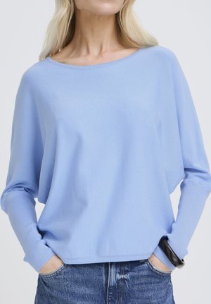 Nő világoskék laza pulóvert visel, hosszú, testhezálló ujjakkal és kék farmert, kezét zsebre téve, fehér háttér előtt.