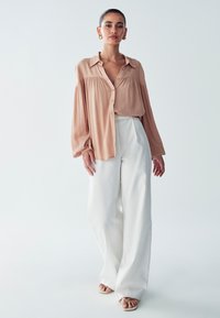 Bežna bluza s gumbi in napihnjenimi rokavi, v kombinaciji s širokimi belimi hlačami. Gladek material, minimalističen dizajn, subtilne drape podrobnosti.
