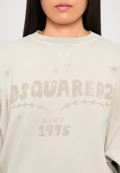 Woman wearing a beige Dsquared2 sweatshirt with faded logo and “Since 1995” text, decorated with subtle star and leaf motifs.
