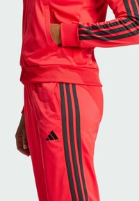 Rode sportieve trainingsoutfit met zwarte driekleurige accenten op de mouwen en zijkanten van de broek. Zachte, gladde textuur en smal ontwerp.