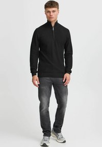 Un pull zippé noir avec un design en tricot texturé, associé à un jean gris et des chaussures de sport grises. Le pull présente un col montant et des poignets côtelés.