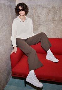 Weißes Langarm-Polo, braune weit geschnittene Hose und weiße High-Top-Sneaker, sitzend auf einer roten Couch vor einem Betonwand-Hintergrund.