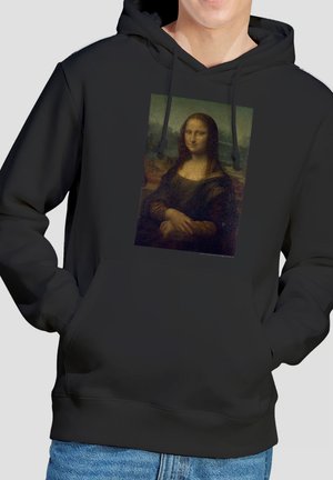 Person trägt einen schwarzen Hoodie mit einem aufgedruckten Mona Lisa-Porträt auf der Vorderseite, Hände in den Taschen des Hoodies, kombiniert mit blauen Jeans.