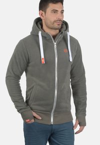 Gray fleece zip-up hoodie with white drawstrings, side pockets, and red brand logo. Soft texture and fitted design.