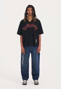 Černá košile s krátkým rukávem a knoflíky s červeným textem "Ed Hardy"; modré denímové cargo kalhoty s bílými výšivkami; černobílé tenisky.