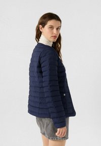 Giubbotto imbottito blu navy con design trapuntato, caratterizzato da una scollatura rotonda e dettagli con bottoni automatici. Indossato sopra un dolcevita chiaro.