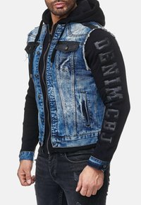 Jeansvest met zwarte mouwen, verstoord blauw stof, schoenen leren accenten, voorkant zakken en "DENIM" tekst op de mouw.