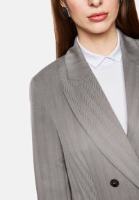 Gray pinstriped blazer with sharp lapels, double-button closure, and textured fabric. White collared shirt visible underneath.