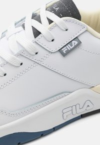 Sneaker in pelle bianca con linguetta in rete grigia e dettagli testurizzati. Presenta lacci bianchi e una suola in gomma blu con il branding Fila.