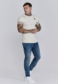 Creme t-shirt met bordeauxstrepen en logo, gecombineerd met blauwe denim slim-fit jeans en witte sneakers. Soepele textuur, casual stijl.