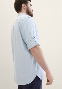 Hellblaues Kurzarmhemd mit umgeschlagenen Ärmeln, strukturiertem Stoff, Knopfakzenten und abgerundetem Saum. Von der Seite getragen.