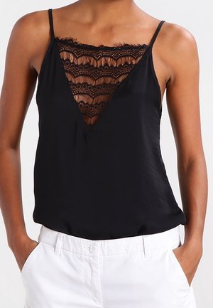 Haut noir sans manches avec empiècement en dentelle à encolure en V, porté avec un pantalon blanc, mains posées décontractées dans les poches, photo cadrée des épaules aux hanches.