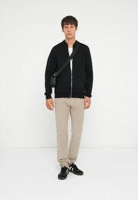 Black zippered bomber jacket, white t-shirt underneath, beige pants, and black sneakers. Small black shoulder bag attached to the jacket.