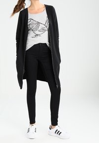 Cardigan noir long sur une chemise grise avec un motif d'oiseau, associé à un jean noir slim et des baskets blanches avec des rayures noires.