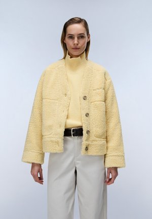 Frau trägt eine hellgelbe Sherpa-Jacke über einem gelben Rollkragenpullover, beige Hose und einen schwarzen Gürtel und steht vor einem schlichten Hintergrund.