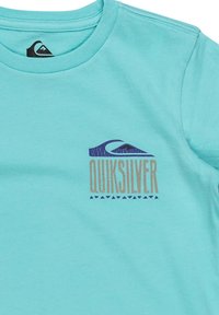 Camiseta turquesa de algodón con mangas cortas, que presenta un estampado gráfico azul y dorado del logo de Quiksilver y un diseño de olas.