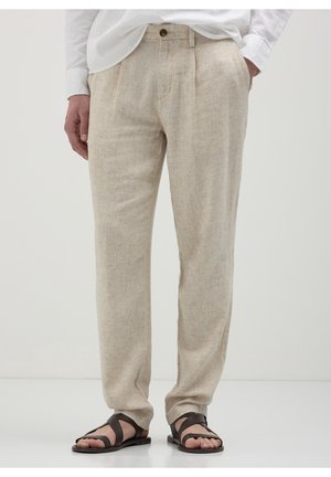 Hombre con pantalones de lino beige con botón y pliegues, combinado con sandalias de cuero marrón oscuro y una camisa blanca.