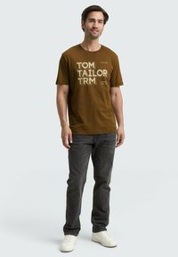 Brun kortærmet t-shirt med hvid trykt tekst. Båret med mørke jeans og lyse sneakers. Casual pasform, bomuldsstof, simpelt design.
