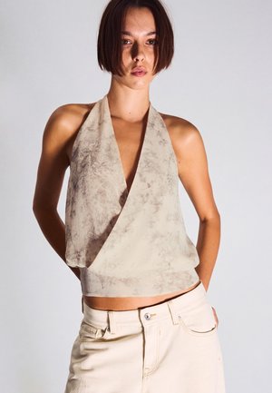 Young woman with short brown hair wears beige tie-dye halter top and cream-colored pants, standing against a plain light background.