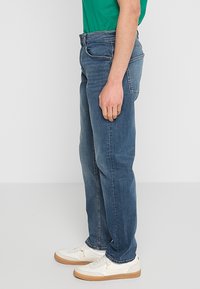 Jeans rectos azules, hechos de mezclilla, con bolsillos delanteros, trabillas para cinturón y un acabado desgastado, combinados con zapatillas blancas.