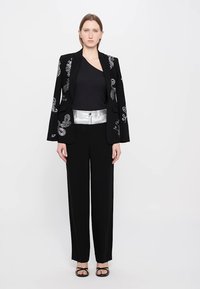 Zwarte blazer met zilveren details, zwarte top, zilveren riem en zwarte wijde broek. Gladde textuur met ingewikkelde patronen op de blazer.