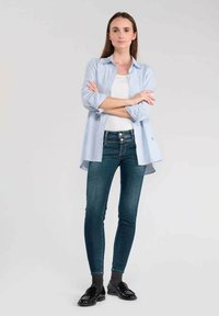 Lichtblauw en wit gestreepte blouse over een witte top, gecombineerd met donkere skinny jeans en zwarte loafers. Glad textiel en een getailleerde pasvorm.