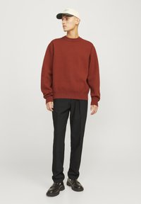 Pull de couleur rouille avec un col rond, associé à un pantalon noir et des mocassins noirs. Le mannequin porte une casquette beige avec une visière sombre.