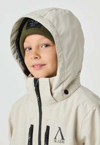 Cream-colored waterproof jacket with a zippered front, black zippers, and two pockets. The hood is lined and a dark green knitted beanie is worn.